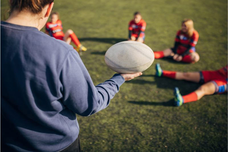 The image shows a person in the foreground holding a white rugby ball and talking to other players who are sitting on the pitch