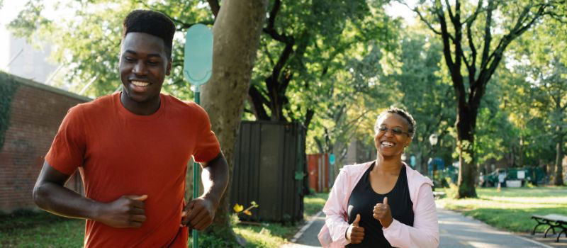 Two people are pictured running along a pathway, between trees, in the sunshine. They are both smiling broadly.