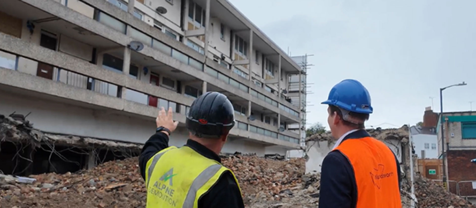 There is a pile of rubble in front of an old building, which is being demolished. Two people in high visibility vests and hart hats are standing in the foreground, looking at the work that is taking place.