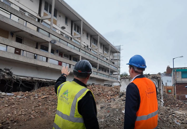 There is a pile of rubble in front of an old building, which is being demolished. Two people in high visibility vests and hart hats are standing in the foreground, looking at the work that is taking place.