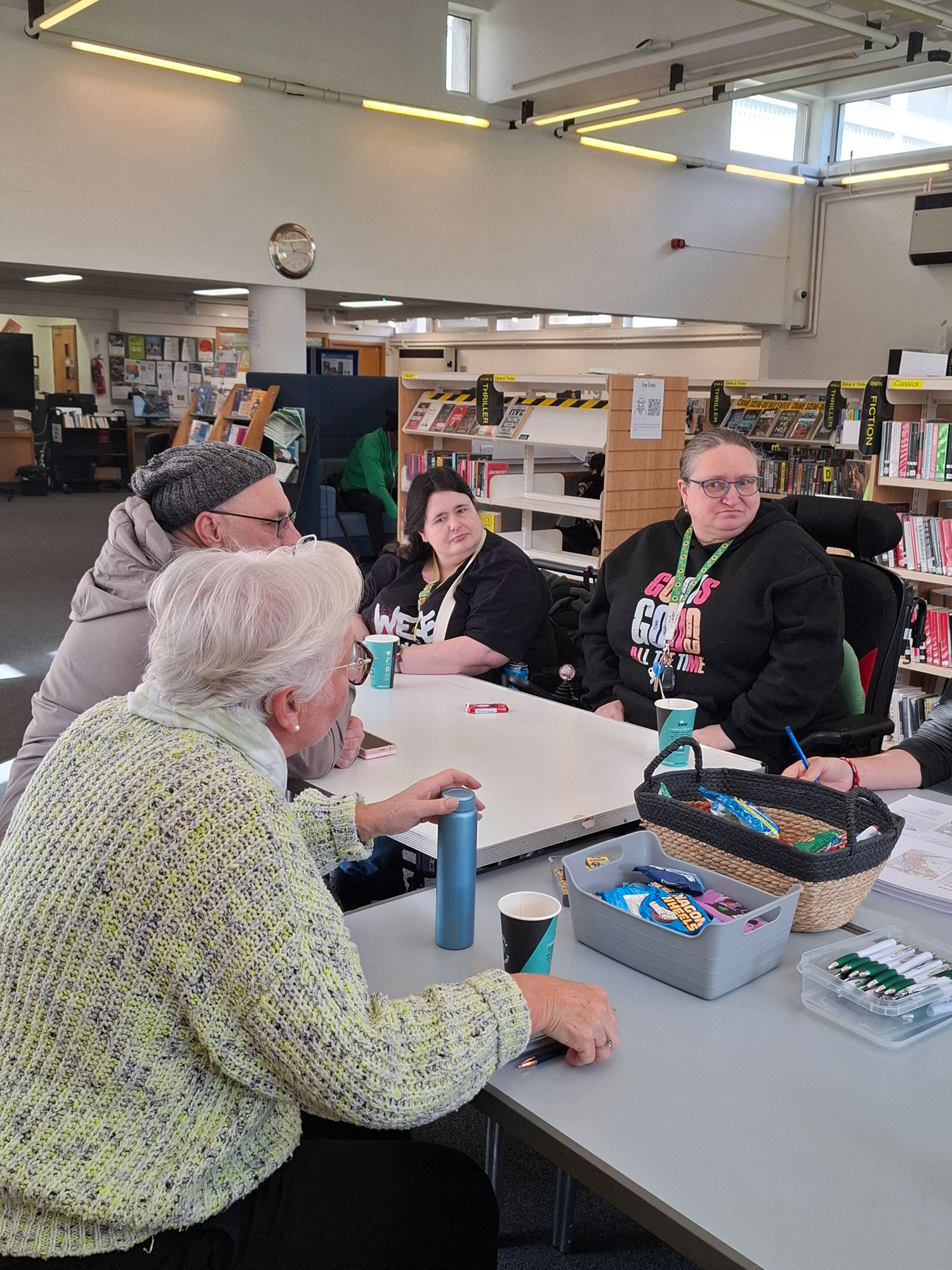 A group of people are featured, sitting around a table in a library, having a discussion.