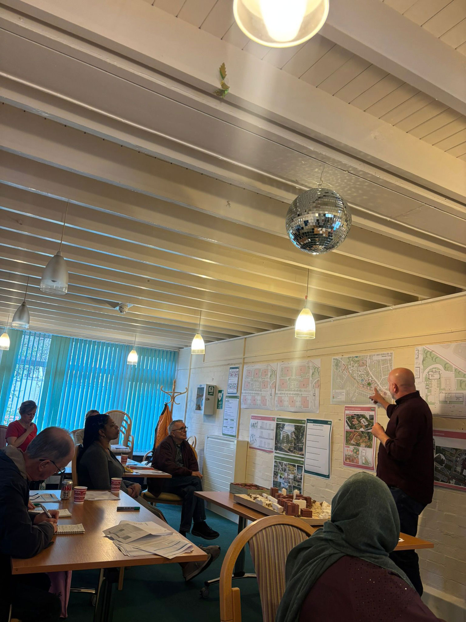 People are pictures sitting at tables looking towards a speaker and taking notes. The speaker is standing, pointing at plans on a wall.
