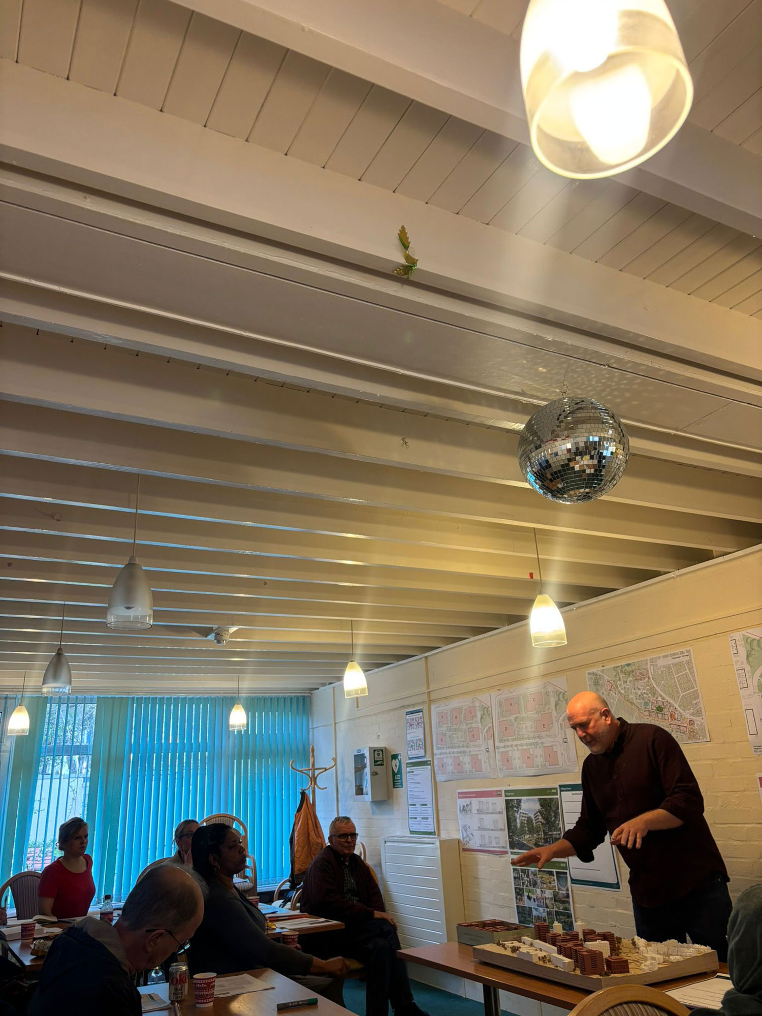 People are pictures sitting at tables looking towards a speaker and taking notes. The speaker is standing, pointing at a model of the estate, which is on a table in front of him.