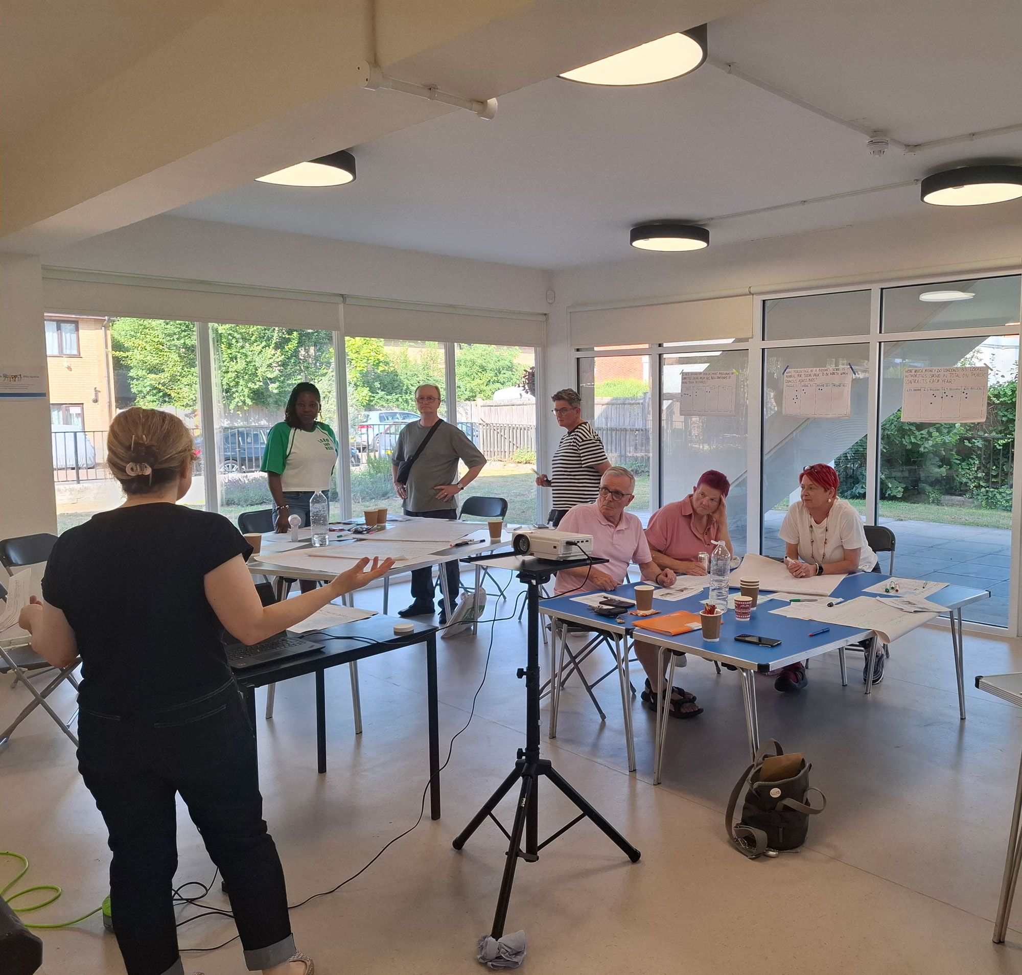 People are pictured sitting and standing around tables, looking towards someone who is standing at the front of the room, speaking.