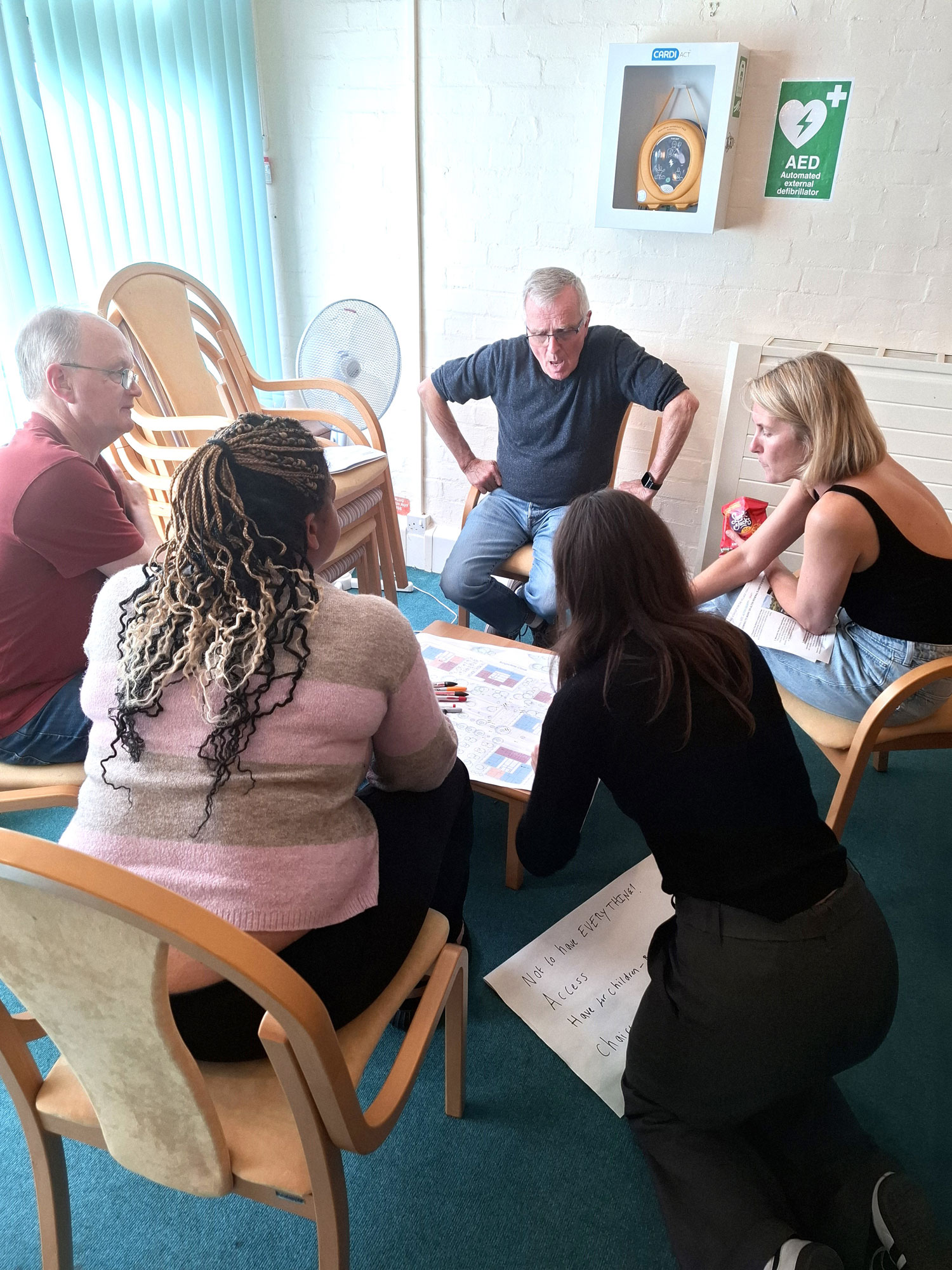 People are pictured sitting around a table having a discussion. The table is covered with plans.