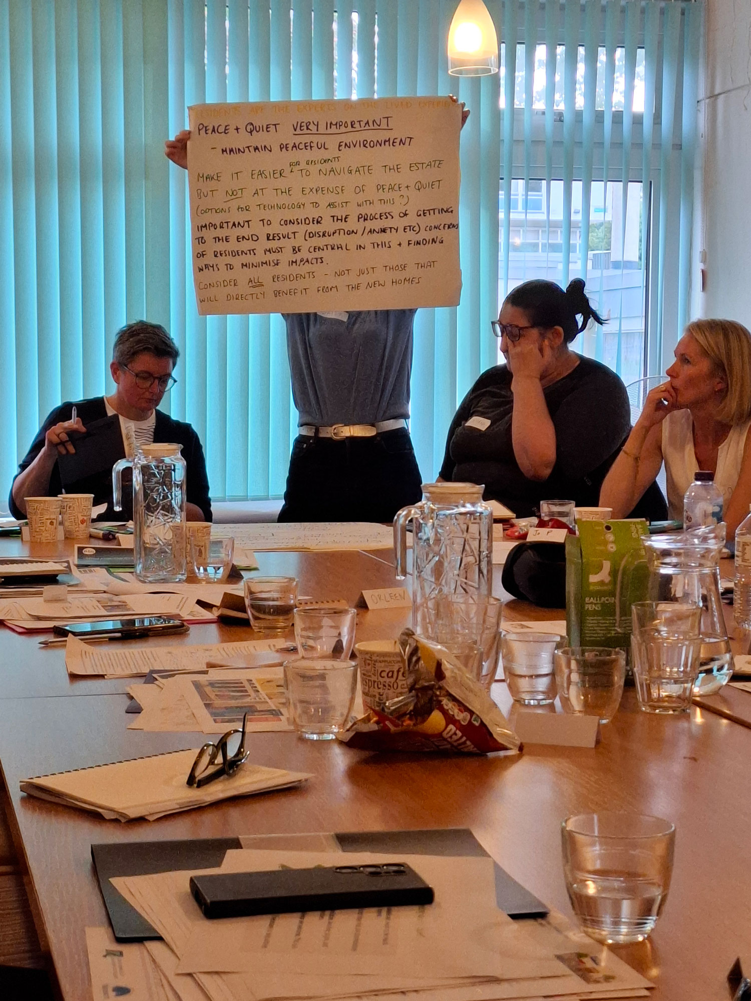 A group of people are sitting around the table. One person is standing up and holding a flipchart with writing on it.