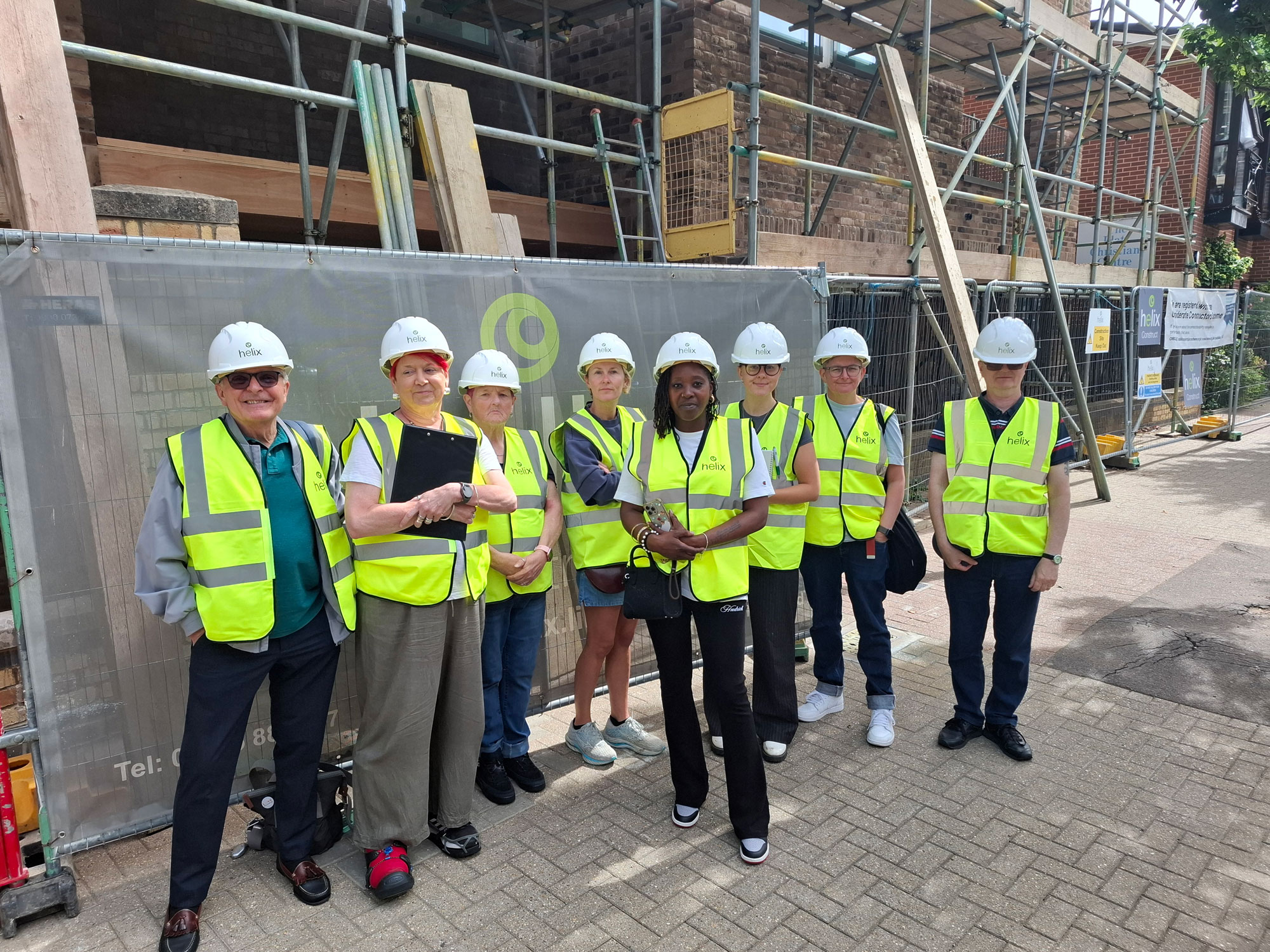 A group of people wearing high visibility vests and hard hats is pictured standing in front of construction barriers and a building that's under construction.