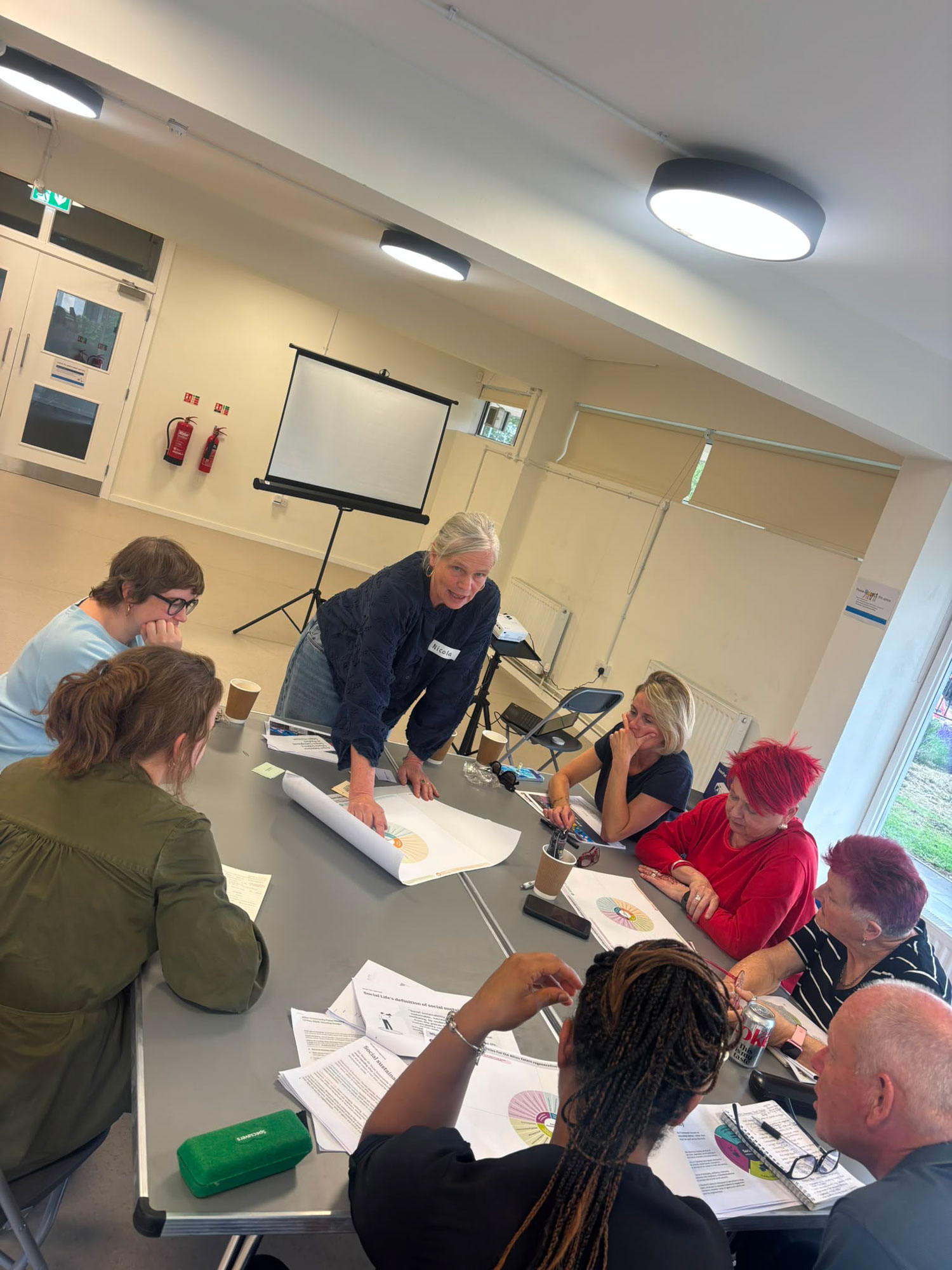 People are sitting around a table listening to someone who is standing at the end of the table, pointing to a chart in the centre of the table.
