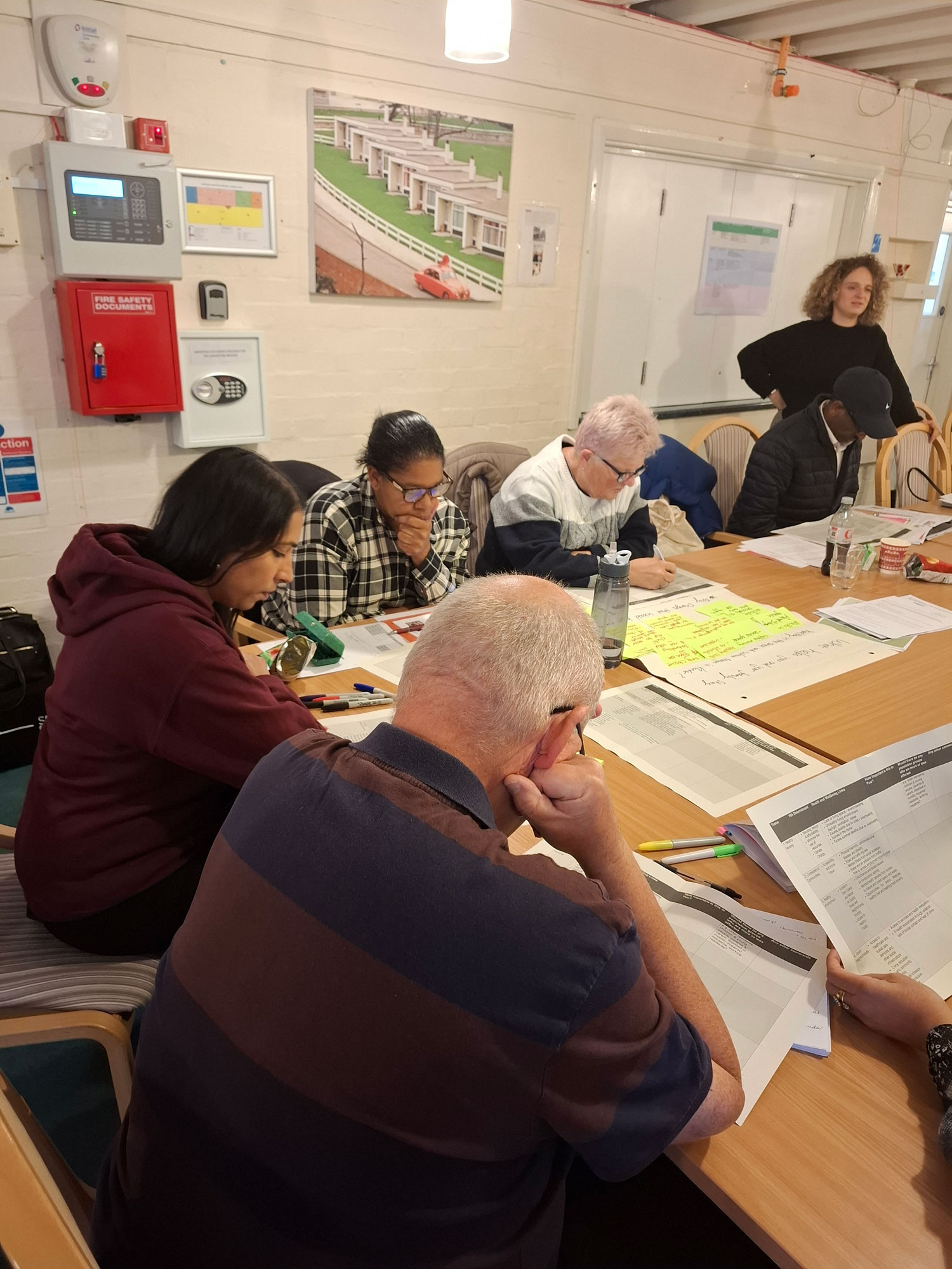 A group of people are sitting and standing around a table, looking thoughtful and making notes.