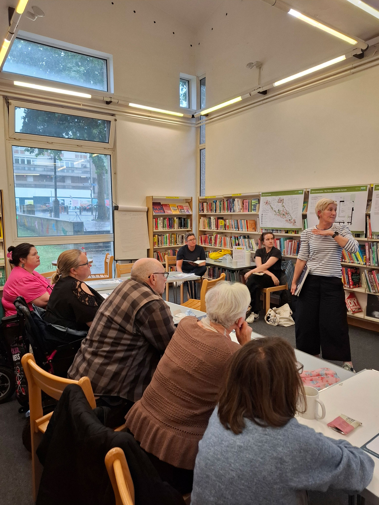 People are pictured sitting in Roehampton Library, looking towards someone who is standing up, making a presentation in front of bookshelves.